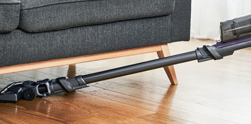Un aspirapolvere senza fili grigio sotto un divano moderno su pavimento di legno.